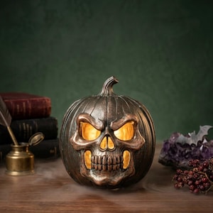 May include: A dark bronze-colored pumpkin decoration featuring a carved skull face on the front. The interior is illuminated with a warm yellow light that shines through the eye sockets and mouth. The pumpkin has a textured, weathered finish.