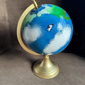 May include: A small globe with a gold-colored base and meridian ring. The globe is painted with blue oceans, green landmasses, and white cloud-like shapes. A small orca figure is visible on the blue ocean. The globe is a decorative item.