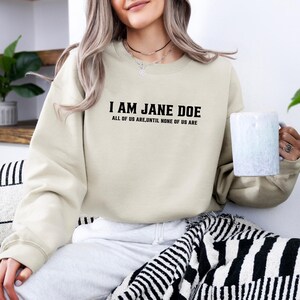 May include: A beige crewneck sweatshirt featuring the black text I AM JANE DOE and the smaller text ALL OF US ARE, UNTIL NONE OF US ARE printed on the chest. The garment has long sleeves with ribbed cuffs and a ribbed neckline, providing a simple design.
