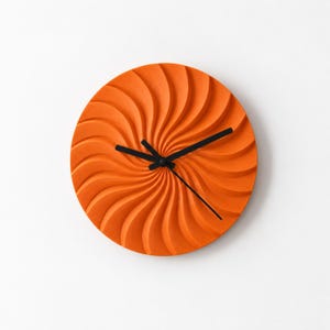 May include: A circular wall clock featuring a vibrant orange face with a three-dimensional, swirling, radial pattern. The clock has three thin, black hands positioned at the centre, indicating the time against the textured, grooved surface.
