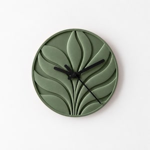 May include: A circular wall clock featuring a sage green face with a raised, three-dimensional relief pattern of radiating, curved leaf-like shapes. The clock has three thin, black hands positioned at the center, indicating the time.