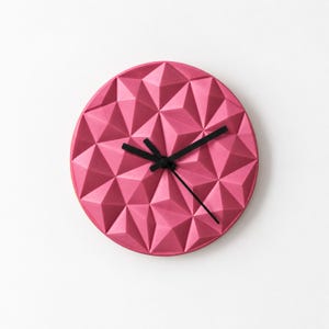 May include: A circular wall clock with a vibrant pink, faceted geometric surface. The clock face features a three-dimensional triangular pattern across its entire front. Three thin, black hands are positioned at the centre of the clock.