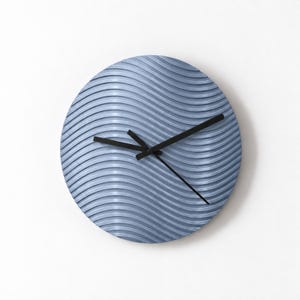 Puede incluir: Un reloj de pared circular con una esfera de color azul claro que presenta un patrón repetitivo de líneas onduladas horizontales. El reloj tiene tres manecillas negras delgadas situadas en el centro, que marcan la hora sobre la superficie texturizada.