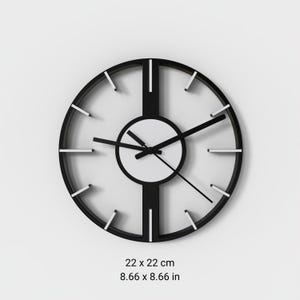 Op de afbeelding: Een ronde wandklok met een zwart frame, een centrale ring en verticale dwarsbalken. De wijzerplaat heeft witte uurmarkeringen en drie zwarte wijzers. De afmetingen onder de klok worden weergegeven als 22 x 22 cm.