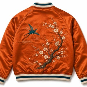 May include: A burnt orange satin bomber jacket featuring intricate embroidery on the back. The design depicts a teal bird in flight alongside a flowering tree branch with white blossoms. The jacket has ribbed cuffs and a collar with teal and white stripes.
