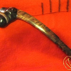 May include: A curved club with a polished black stone head featuring a silver-toned band at the neck. The handle is made of dark, variegated wood with a visible grain pattern. A thin yellow cord is attached to the end of the handle.