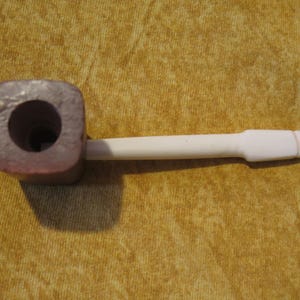 May include: A smoking pipe featuring a square-shaped, dark reddish-brown bowl with a circular opening at the top and a long, smooth, white stem extending from the side. The pipe is constructed with a distinct contrast between the dark bowl and the light stem.