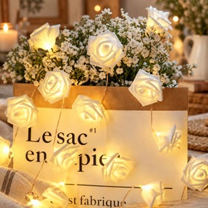 Op de afbeelding: Een lichtsnoer met LED-lampjes in de vorm van witte rozen die een warme gloed verspreiden. De lampjes zijn gedrapeerd over een rechthoekige papieren zak met de tekst Le sac en papier #1 st fabrique in zwarte letters op de voorzijde.
