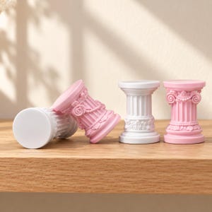 May include: Four small decorative columns in pastel pink and white. Two columns are standing upright, while two are lying on their sides. Each column features intricate relief details, including fluted shafts and ornate floral scrollwork at the base.