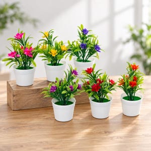 May include: Six small artificial potted plants in white plastic pots. The plants feature green foliage and vibrant flowers in shades of pink, yellow, purple, and orange. Each plant is arranged in a simple, clean white container with green base.