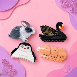 May include: Four animal-shaped hair clips: a gray and white rabbit, a black swan with gold wing details and an orange beak, a white and black penguin with the text Barrette La France, and a peach-colored piece featuring three small bird figures.