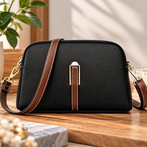 May include: A black, dome-shaped crossbody bag with a textured finish, featuring a brown leather strap with white stitching and a central vertical brown leather accent strip secured by a gold-toned metal clasp hardware.