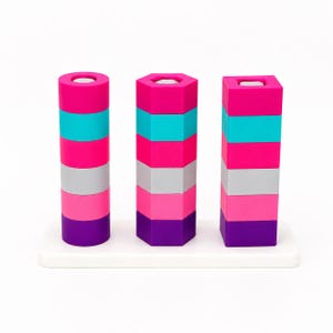 May include: A stacking toy featuring three vertical columns on a white rectangular base. Each column consists of five stacked geometric shapes in shades of hot pink, turquoise, light pink, gray, and purple, with circular, hexagonal, and square profiles.