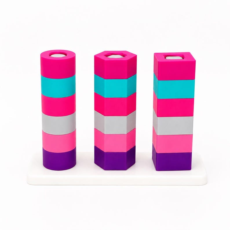 May include: A stacking toy featuring three vertical columns on a white rectangular base. Each column consists of five stacked geometric shapes in shades of hot pink, turquoise, light pink, gray, and purple, with circular, hexagonal, and square profiles.
