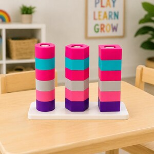 May include: A stacking toy featuring three vertical columns of colorful blocks on a white rectangular base. Each column consists of six blocks in shades of pink, teal, grey, and purple. The columns have circular, hexagonal, and square shapes.