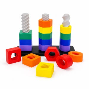 Op de afbeelding: Een speelgoedset met drie verticale schroefdraadpalen op een zwarte basis, met diverse kleurrijke geometrische vormen zoals vierkanten, zeshoeken en cirkels die op de palen gedraaid kunnen worden, samen met verschillende losse onderdelen.