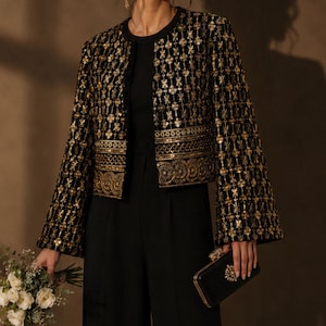 May include: A black jacket featuring intricate gold embroidery and sequin detailing in a floral grid pattern, paired with black trousers and a black clutch with gold trim. The jacket has wide sleeves and a cropped length with a decorative border.