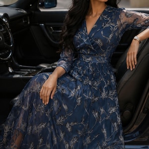 May include: A navy blue midi dress featuring a V-neckline, three-quarter length sleeves, and a sheer overlay with a metallic silver leaf pattern. The dress has a fitted waist and a flared skirt, paired with black strappy high-heeled sandals.