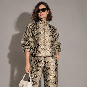 Peut inclure: Un ensemble assorti composé d'une veste zippée à col montant et manches longues et d'un pantalon large, tous deux dotés d'un imprimé peau de serpent beige, noir et crème. La tenue est complétée par des baskets blanches et un petit sac à main blanc.
