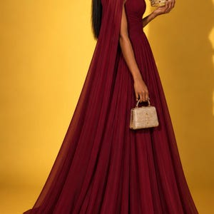 May include: A floor-length burgundy gown with a high neckline and sheer cape sleeves, paired with a small gold structured handbag and a gold crown featuring red stones. The dress has a pleated bodice and a flowing, floor-length skirt.