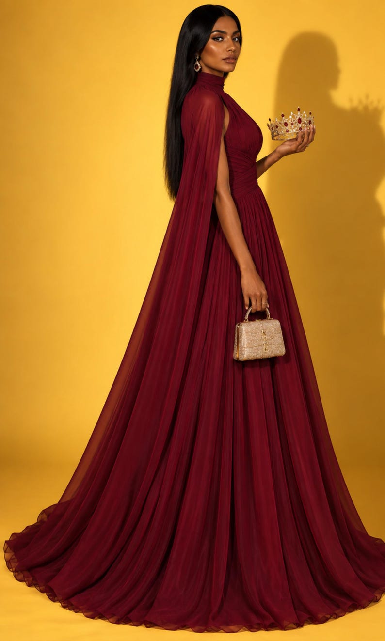 May include: A floor-length burgundy gown with a high neckline and sheer cape sleeves, paired with a small gold structured handbag and a gold crown featuring red stones. The dress has a pleated bodice and a flowing, floor-length skirt.