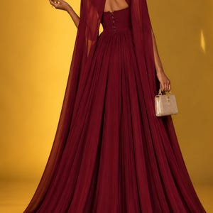 May include: A floor-length burgundy gown featuring a sheer cape overlay, a high neckline with a keyhole cutout, and a row of buttons along the bodice. The dress has a full, pleated skirt and is paired with a gold crown and a small gold handbag.