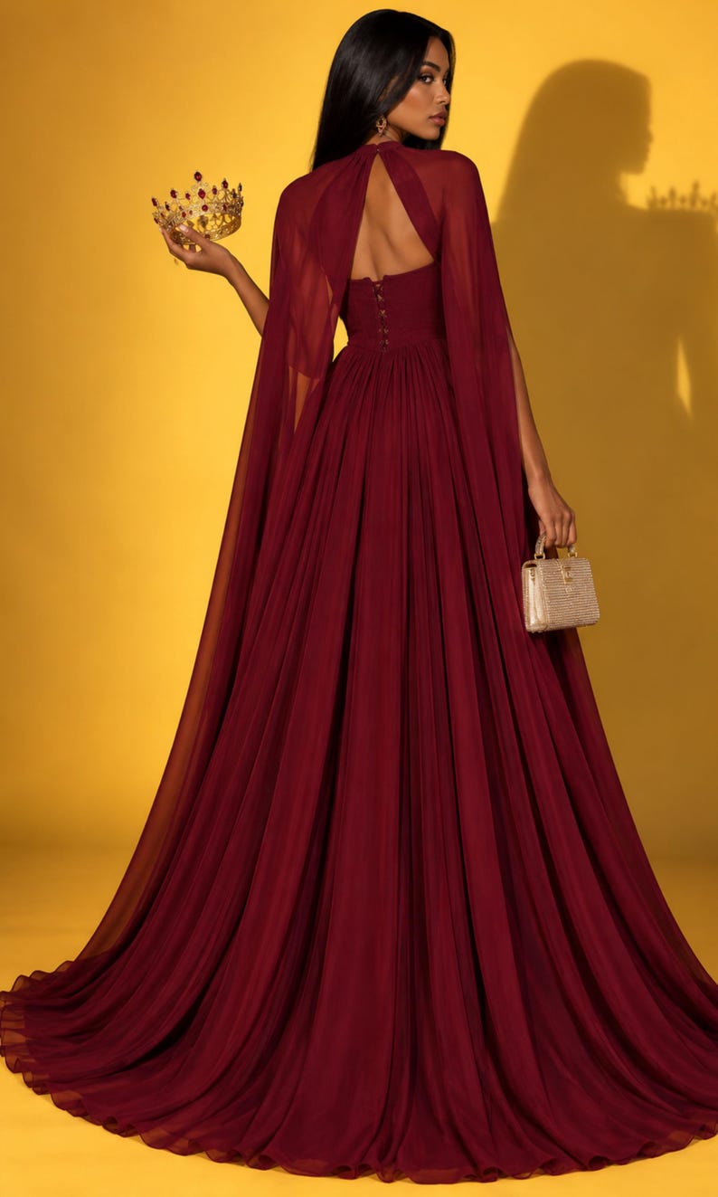 May include: A floor-length burgundy gown featuring a sheer cape overlay, a high neckline with a keyhole cutout, and a row of buttons along the bodice. The dress has a full, pleated skirt and is paired with a gold crown and a small gold handbag.