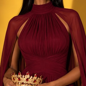 May include: A floor-length burgundy gown with a high neckline, gathered bodice, and attached cape sleeves. The garment is paired with a gold crown featuring red oval stones and a small gold handbag with a textured surface and a matching red stone.
