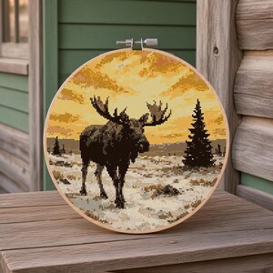 May include: A circular wooden embroidery hoop containing a cross-stitch pattern of a dark brown moose standing in a snowy landscape. The background features a vibrant yellow and orange sunset sky with a single dark evergreen tree silhouette.