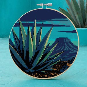 Puede incluir: Un bastidor de bordado circular que contiene un patrón de punto de cruz de una planta de agave grande en primer plano con una silueta de montaña oscura al fondo. La paleta de colores consta de tonos azul marino, turquesa, verde claro y marrón.