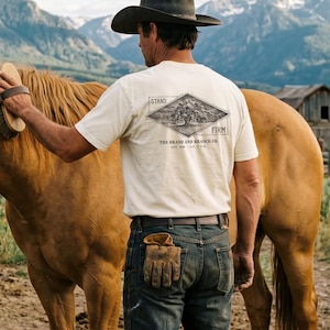 May include: A cream-colored short-sleeve t-shirt featuring a black graphic on the back with a tree inside a diamond shape, the text STAND FIRM, THE BRAND AND BRANCH CO., and EST. 2026 - LA U.S.A. The shirt is worn with dark blue denim jeans.