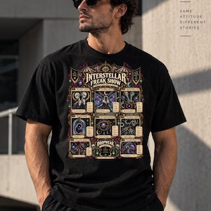 May include: A black short-sleeved t-shirt featuring a colourful graphic print titled Interstellar Freak Show. The design includes multiple framed illustrations of cosmic entities and celestial elements with detailed text and ornate borders.