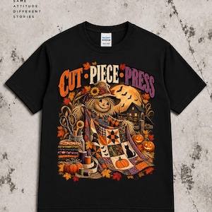 Può includere: Una t-shirt nera a maniche corte con una stampa grafica di uno spaventapasseri che tiene in mano una trapunta patchwork colorata. Il testo recita CUT PIECE PRESS. Il design include foglie autunnali, zucche, un gatto nero, forbici e una luna piena sullo sfondo.