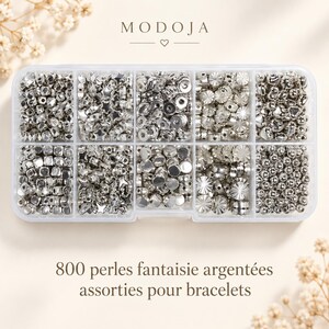 Peut inclure: Une boîte de rangement en plastique transparent à dix compartiments contenant 800 perles en métal argenté de formes variées, incluant des étoiles, des cubes, des sphères et des disques texturés. Le texte MODOJA et 800 perles fantaisie argentées assorties pour bracelets sont visibles.