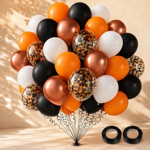Peut inclure: Un grand bouquet de ballons noirs, blancs, orange et cuivrés métallisés, incluant plusieurs ballons transparents remplis de confettis noirs et orange. Les ballons sont attachés avec des rubans noirs fins, et deux bobines de ruban noir sont posées en dessous.