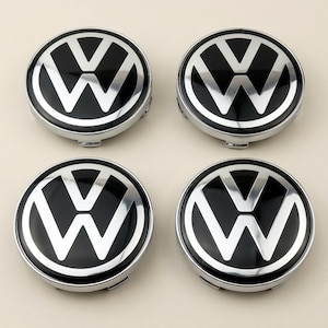 May include: A set of four circular wheel center caps featuring the Volkswagen logo in silver against a glossy black background. Each cap has a polished chrome-colored outer ring and a raised V and W emblem design with a smooth, reflective finish.
