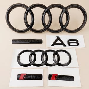 Peut inclure: Un ensemble d'emblèmes automobiles noir brillant comprenant un grand logo Audi à quatre anneaux, un logo plus petit, un badge A6, un badge quattro et deux badges S-line avec des accents rouges et noirs, présentés sur du papier support blanc.