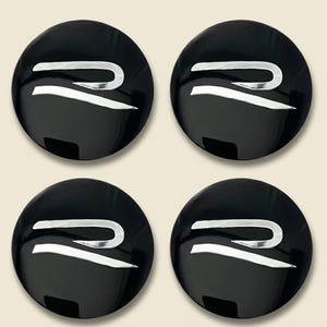 Könnte beinhalten: Ein Set aus vier runden Radnabendeckeln mit glänzend schwarzer Oberfläche und einem markanten, silberfarbenen Buchstaben R als Emblem in der Mitte. Dieses Autozubehör zeichnet sich durch eine glatte, reflektierende und abgerundete Form aus.