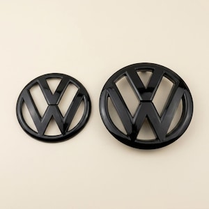 Puede incluir: Dos emblemas circulares negros con el logotipo de Volkswagen, que consiste en una V y una W estilizadas dentro de un círculo. Los emblemas tienen un acabado brillante y se presentan en dos tamaños diferentes, siendo uno más grande que el otro.
