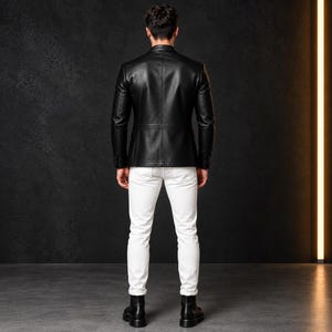 Full-length shot of a man from behind, wearing a black leather blazer, white jeans, and black boots against a dark backdrop.