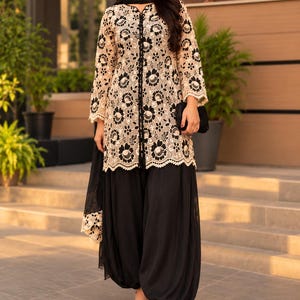 May include: A long-sleeved tunic featuring a beige lace overlay with a black floral pattern, paired with solid black loose-fitting trousers and a matching black scarf. The tunic has a central black button placket and a scalloped hemline.