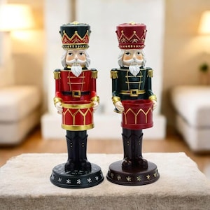 Puede incluir: Dos figuras de cascanueces sobre bases circulares. La figura de la izquierda viste chaqueta roja y sombrero verde, mientras que la de la derecha viste chaqueta verde y sombrero rojo. Ambas sostienen un recipiente con forma de tambor y detalles dorados.