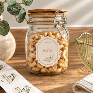 May include: A clear glass storage jar with a bamboo lid and a metal wire bail closure, filled with cashew nuts. A cream-coloured rectangular label on the front features the text PANTRY and NUTS with a botanical line drawing illustration.