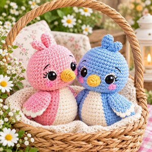 May include: Two crocheted bird plushies, one pink and one blue, each featuring a cream-coloured belly, a small yellow beak, large black safety eyes with long eyelashes, and pink blush spots on their cheeks, sitting inside a woven wicker basket.