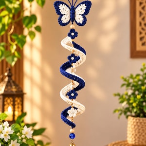May include: A hanging crochet ornament featuring a dark blue and white butterfly at the top, followed by a spiraling dual-colored blue and white ribbon structure accented with small crochet flowers, wooden beads, and a gold-toned metal bell at the base.
