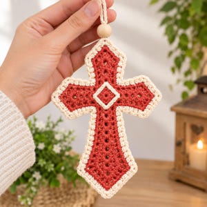 May include: A decorative hanging cross ornament crafted from crochet yarn in a deep rust-red color with a cream-colored border. The cross features a central diamond motif and is suspended by a matching cream yarn loop with a single wooden bead.