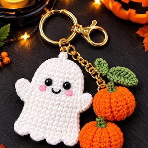 May include: A crochet keychain featuring a white ghost with black eyes and pink cheeks, attached to a gold-toned metal ring. The keychain includes two small orange pumpkins with green stems and a single green leaf, all crafted from yarn.
