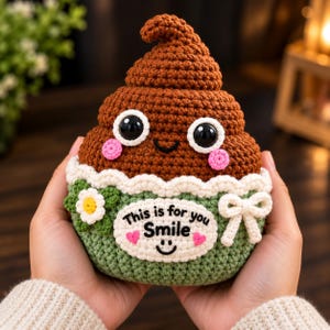 May include: A brown crocheted plush toy shaped like a swirl with large black eyes, pink cheeks, and a green base. It features a white scalloped trim, a small white flower, a white bow, and a white patch with the text This is for you Smile.