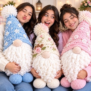 May include: Three handmade crochet gnomes featuring tall pointed hats with pom-poms. The left gnome has a blue hat with white flower accents, the center gnome has a cream hat with floral embroidery, and the right gnome has a pink hat with white flowers.