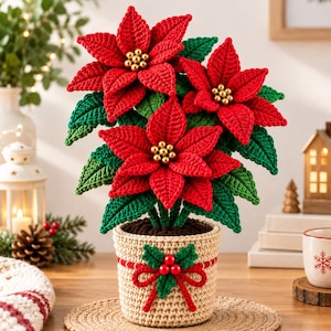 Puede incluir: Una planta de flor de Pascua tejida a ganchillo con pétalos rojos vibrantes y hojas verdes, con centros de cuentas doradas. La planta está en una maceta tejida de color beige decorada con un lazo de lana roja, hojas de acebo y tres cuentas rojas.